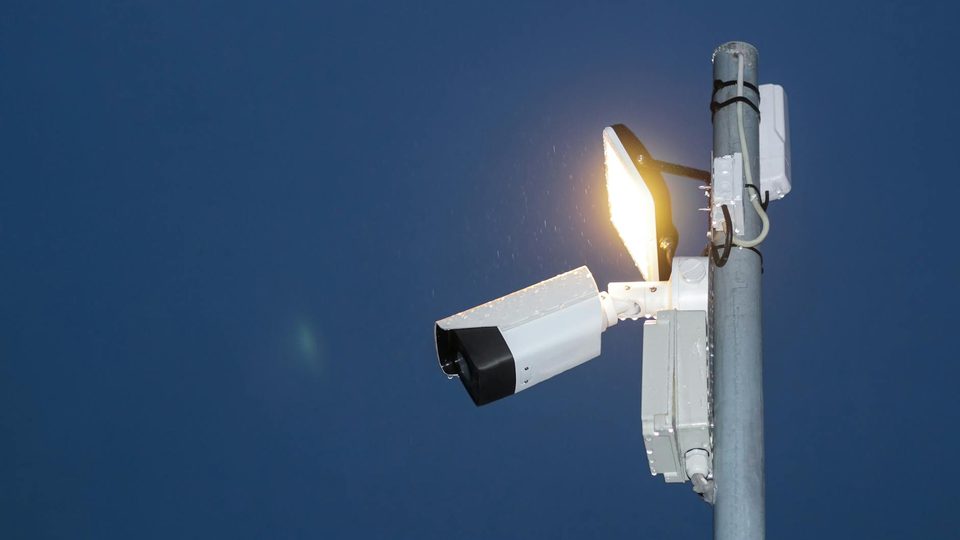 A surveillance camera and exterior light mounted against a blue sky.